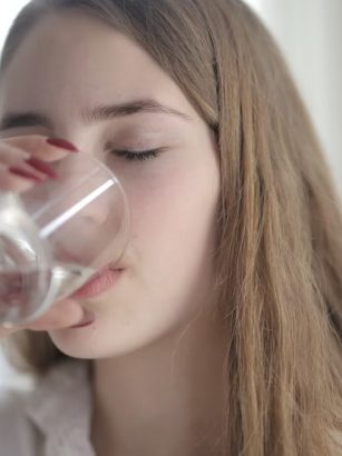A young woman with brown hair drinks water from a clear glass indoors, eyes closed.
