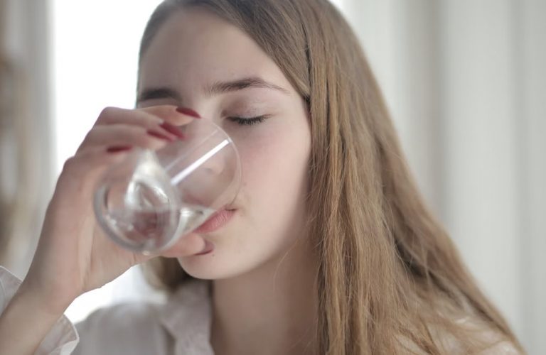 A young woman with brown hair drinks water from a clear glass indoors, eyes closed.